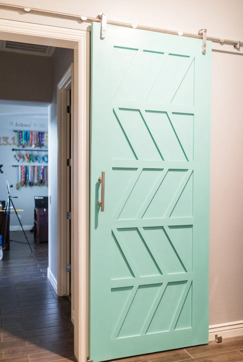 A mint green sliding barn door with diagonal and horizontal cut-out patterns, mounted on a metal track, partially open to reveal a room with dark wood flooring and some furniture.