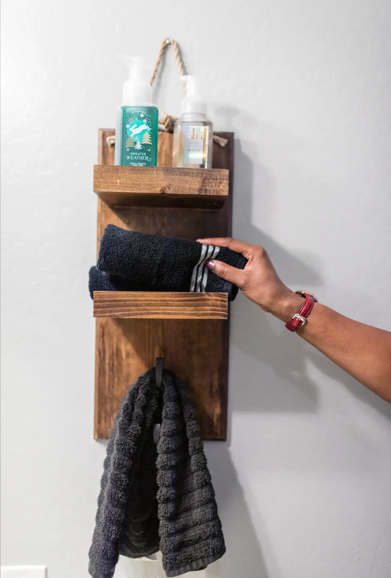 A wooden wall-mounted organizer with three compartments holding hand sanitizer and a towel, with a hand reaching to place a folded towel in the middle compartment.
