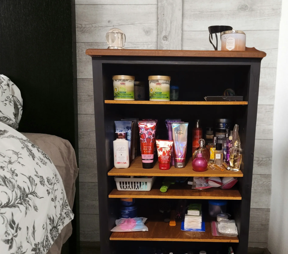 A black bookshelf with five wooden shelves filled with various personal care and beauty products, including lotions, perfumes, and containers, placed next to a bed with floral-patterned bedding in a room with light gray wooden wall panels.