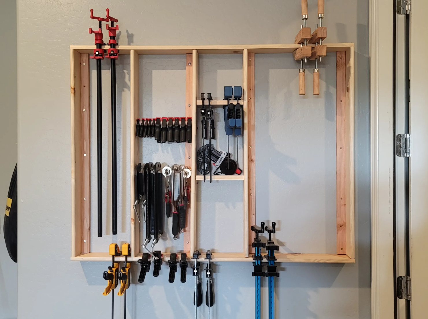 A wooden wall organizer with various clamps, hand tools, and accessories for woodworking or DIY projects. The organizer has multiple sections and hooks for different tools.
