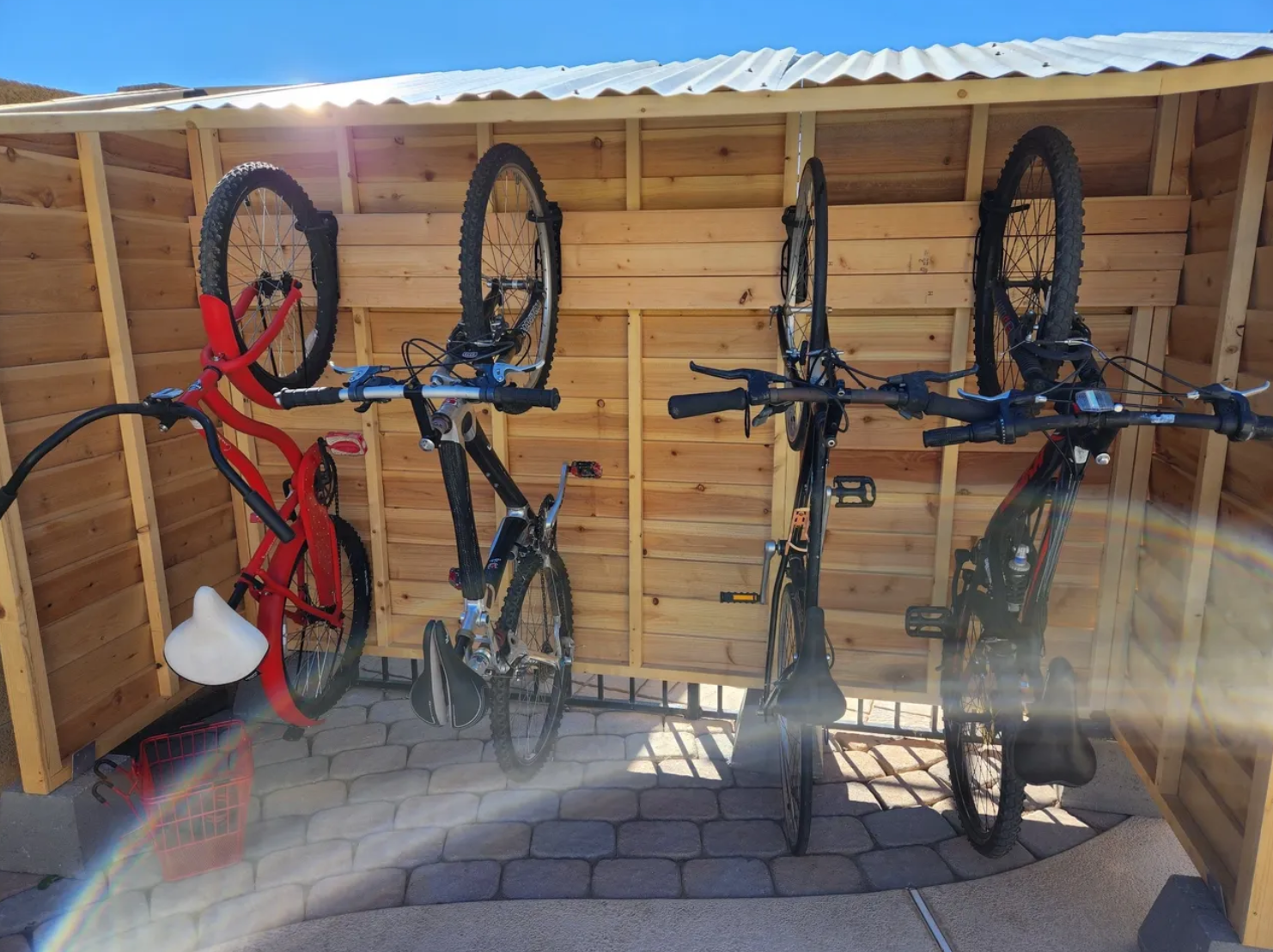 Four bicycles mounted on a wooden bike rack with a small red basket and helmet holder at the bottom left, set against a light wooden fence under a metal roof, with sunlight creating a rainbow flare in the bottom left corner.