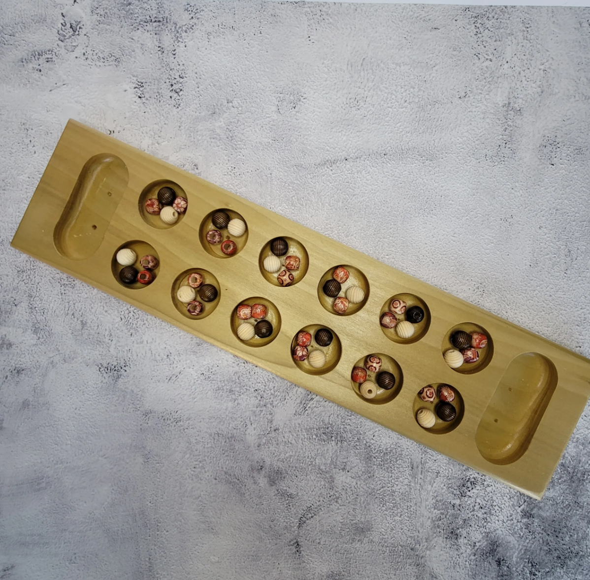 A wooden game board with multiple circular indentations filled with marbles of various colors and patterns, on a textured white surface.
