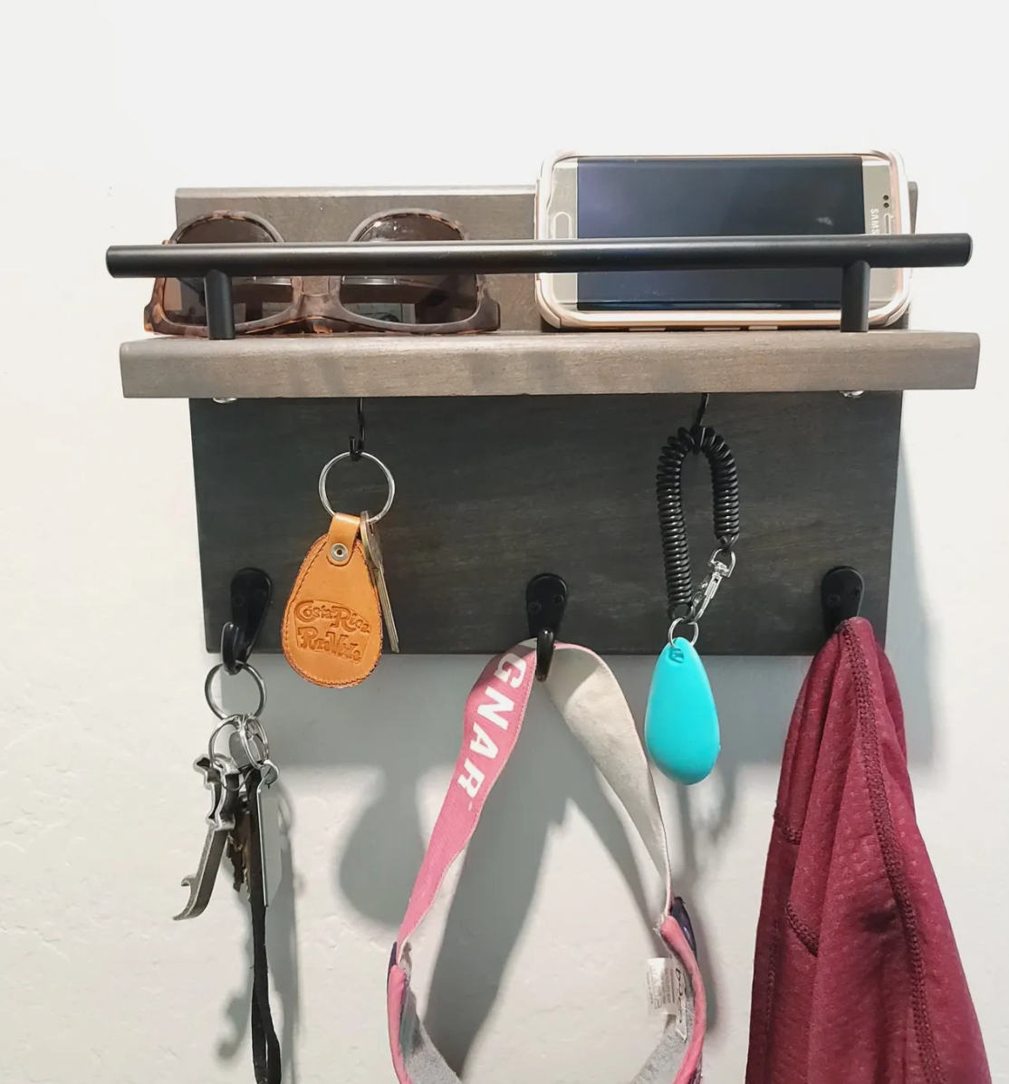 Wall-mounted organization station with a shelf holding sunglasses and a smartphone, hooks holding keys, a leather tag, a pink leash, a blue pet toy, and a red cloth.
