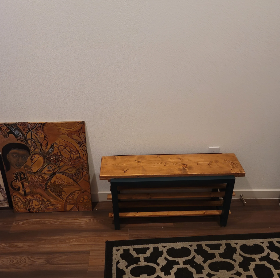 A wooden bench with a black metal frame, a large piece of artwork on the floor showing an abstract portrait of a person with detailed background, a patterned rug on a wooden floor, plain white wall in the background.