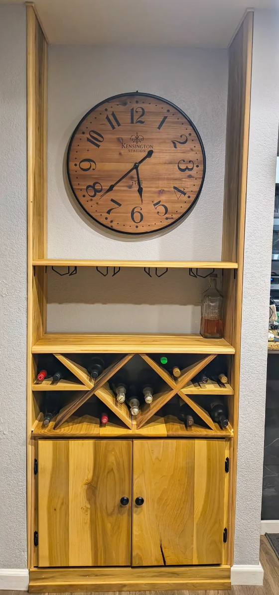 A wooden wine cabinet with a large clock above it. The cabinet has a shelf, a wine rack with eight bottles, and closed storage with black handles. There is also a bottle of liquor on the middle shelf.