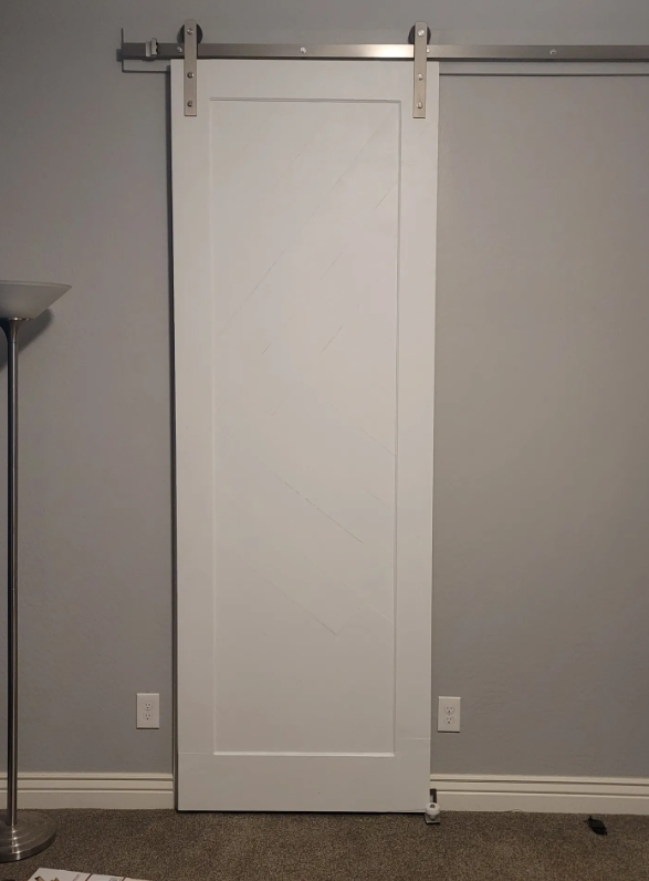 A white sliding door on a metal track, mounted on a gray wall, with a floor lamp on the left and electrical outlets near the floor.