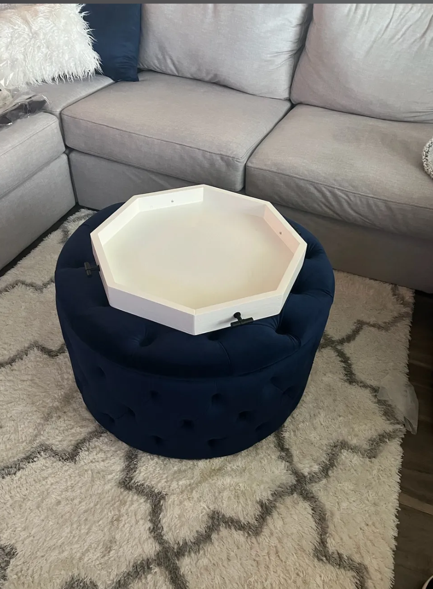Empty white octagonal tray on a round navy tufted ottoman in front of a gray sectional sofa with pillows, on a beige patterned rug.