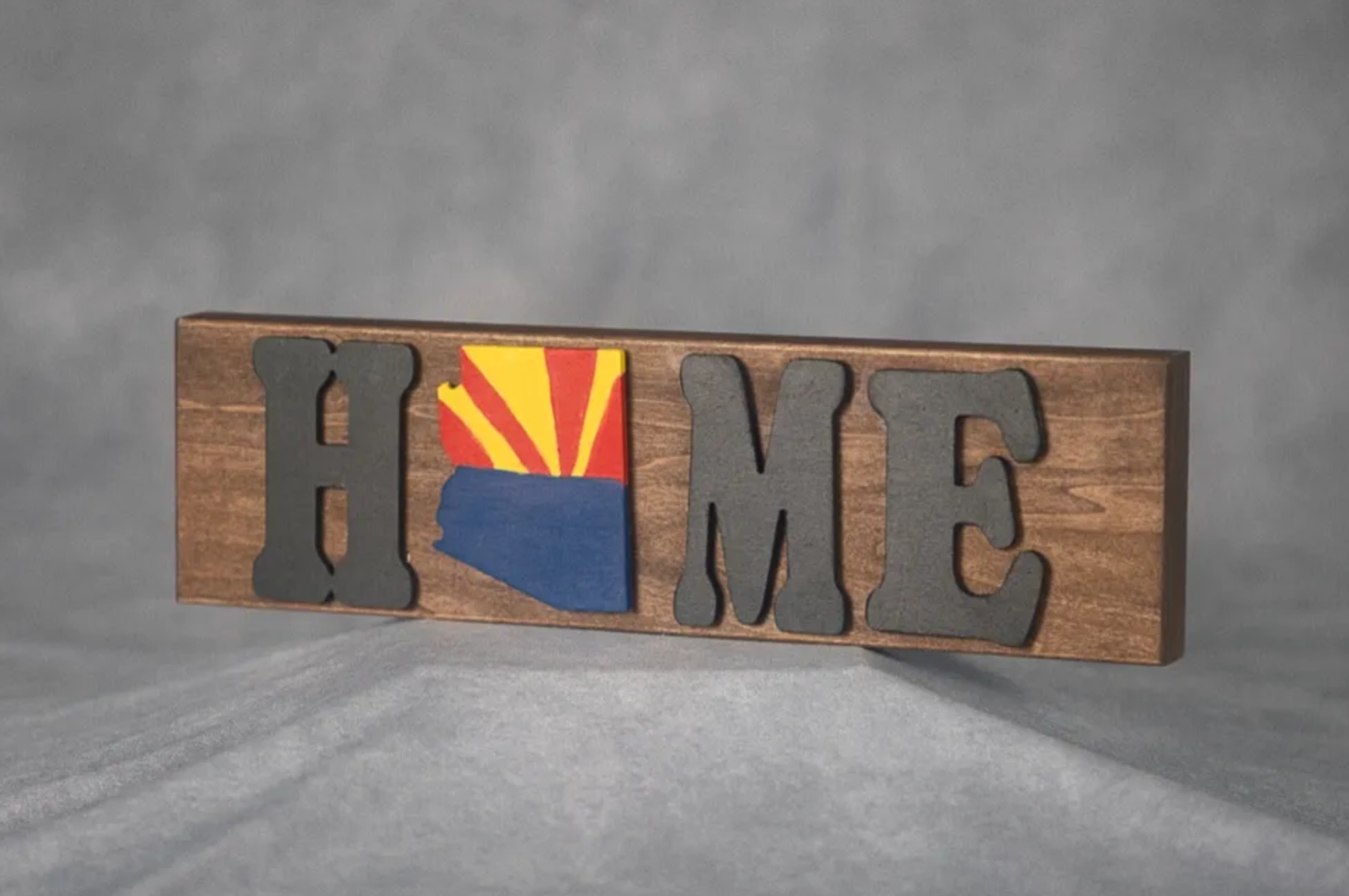 Wooden decorative sign spelling "HOME" with the letter "O" replaced by a painted Arizona state outline featuring the state flag design.