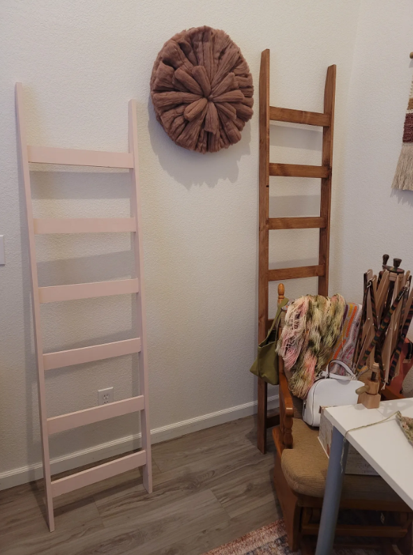 Two wooden ladders leaning against a wall, with a decorative round textile wall hanging above them, and some chairs and textiles in the foreground.