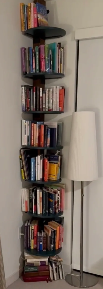 A spiral bookshelf filled with various books next to a white floor lamp in a corner of a room.