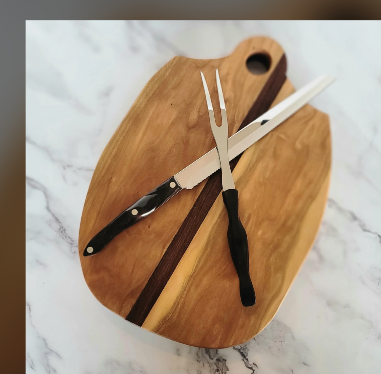 A wooden cutting board with a black-handled serrated bread knife and a carving fork placed on top, on a white marble surface.