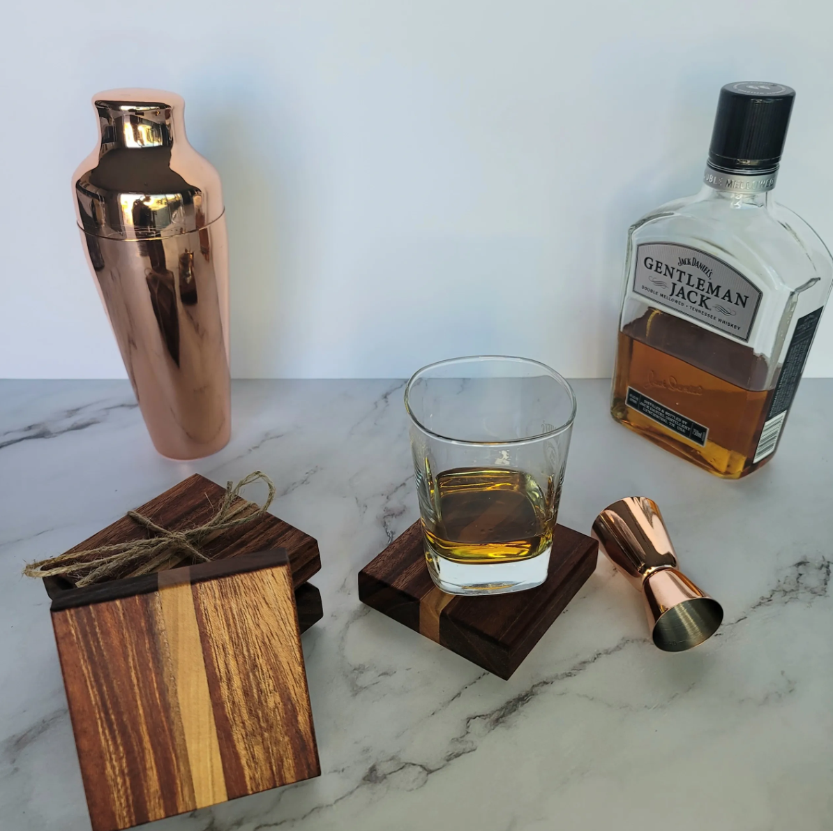 A marble countertop with a pink metallic cocktail shaker, a bottle of Jack Daniel's Gentleman Jack whiskey, a glass of whiskey on a wooden coaster, a cocktail jigger, and a small wooden block with twine.