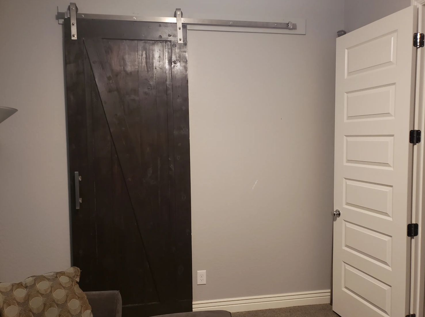 Interior view of a room with a dark sliding barn door on metal rails, a white paneled door, a beige patterned cushion, and a small shelf on the wall.