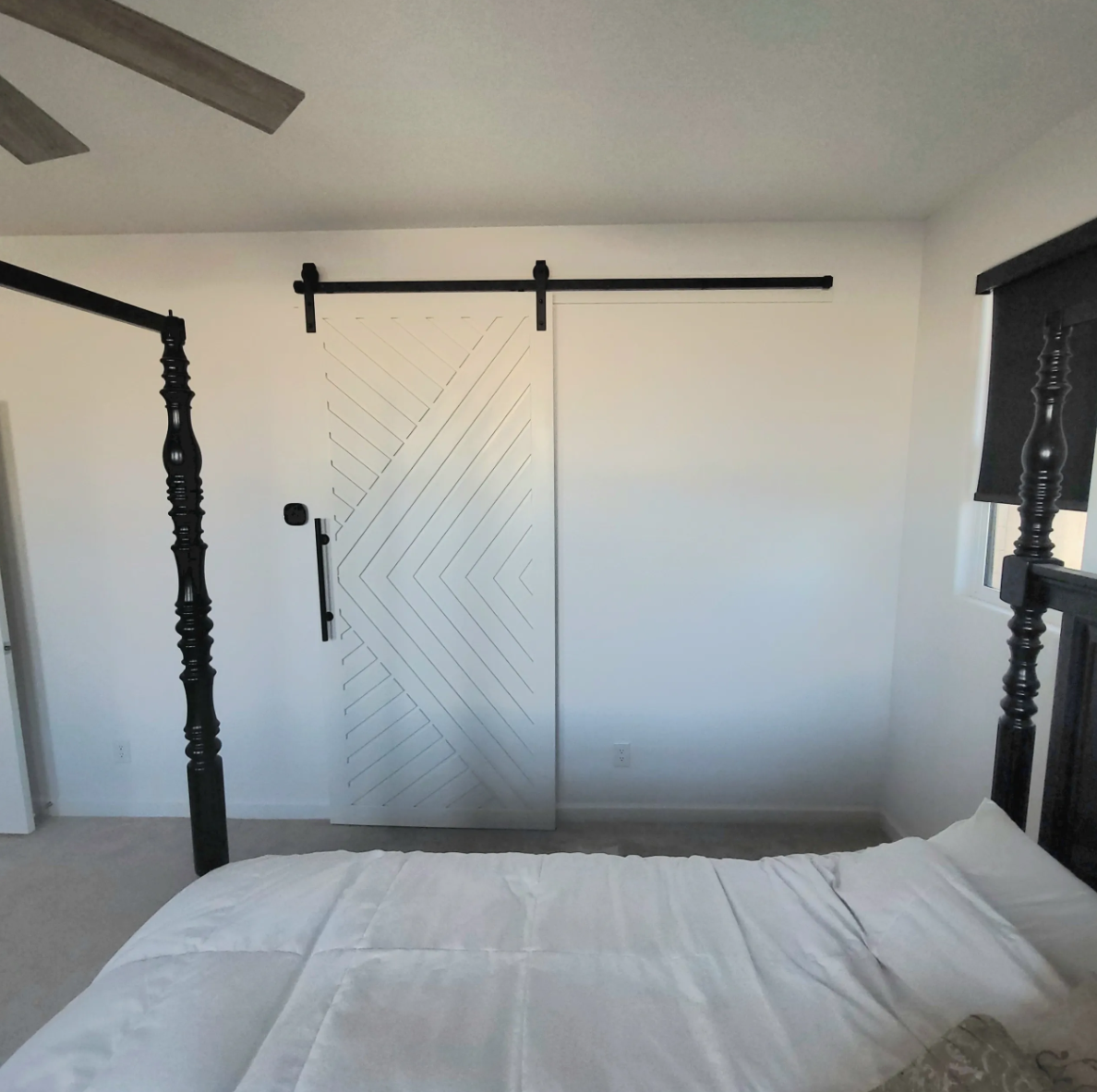 A bedroom with a four-poster bed, white bedding, a white door with geometric design, sliding barn door hardware, and a window with a dark shade.