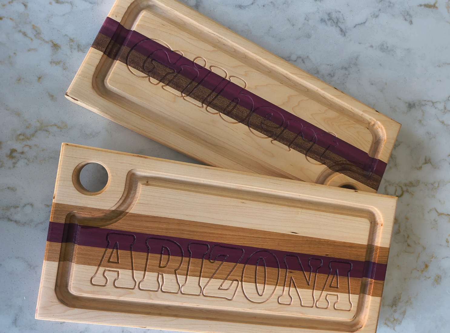 Two wooden ARIZONA-themed cutting boards with engraved text, placed on a marble surface. One board has a handle with a hole, both boards feature a purple stripe and carved letters.
