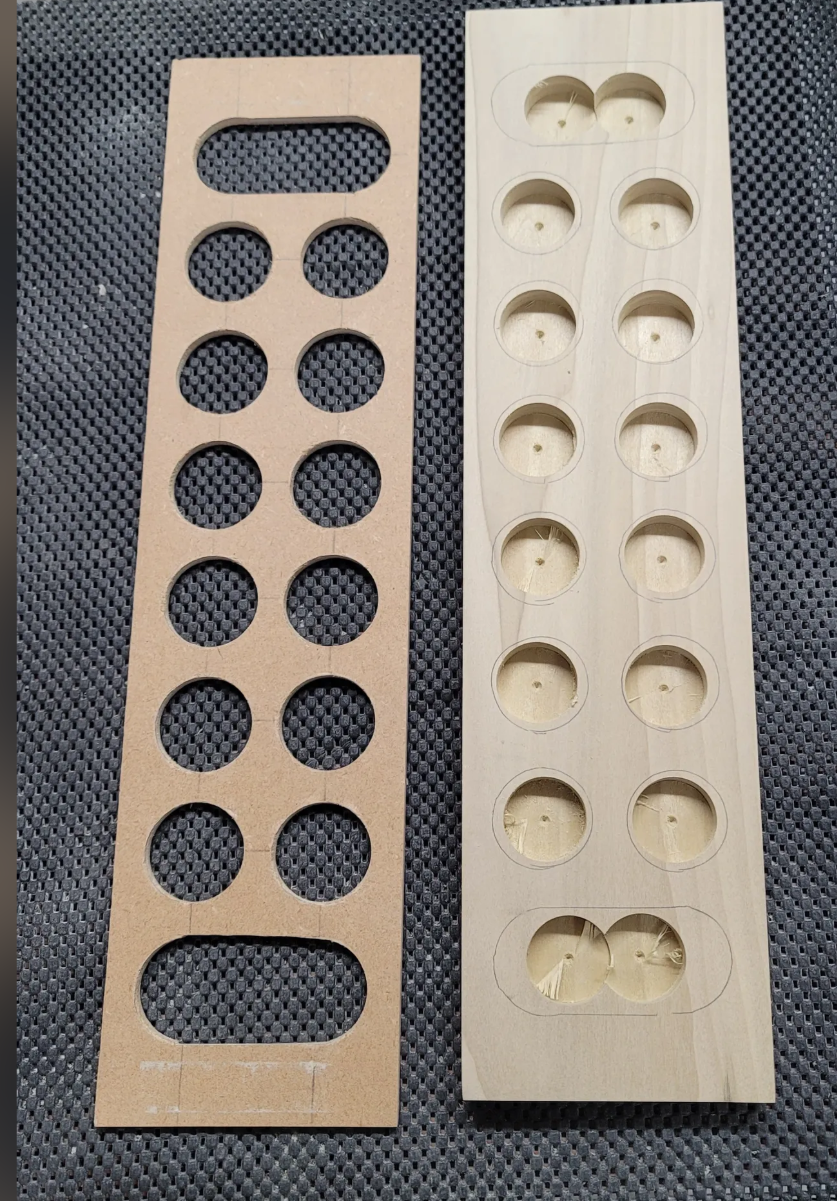 A template for a mancala game next to a strip of wood with circular and elongated holes, likely for a woodworking or crafting project, placed on a textured black surface.