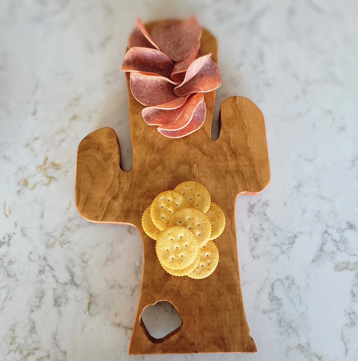 A wooden cactus-shaped tray with a pink succulent plant and round yellow crackers on it, placed on a marble surface.