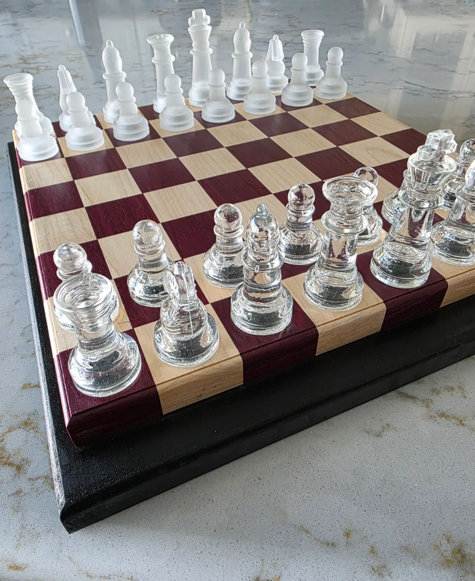 A chessboard with frosted and glass chess pieces set up for a game on a wooden board with squares made of purple heart and maple woods.