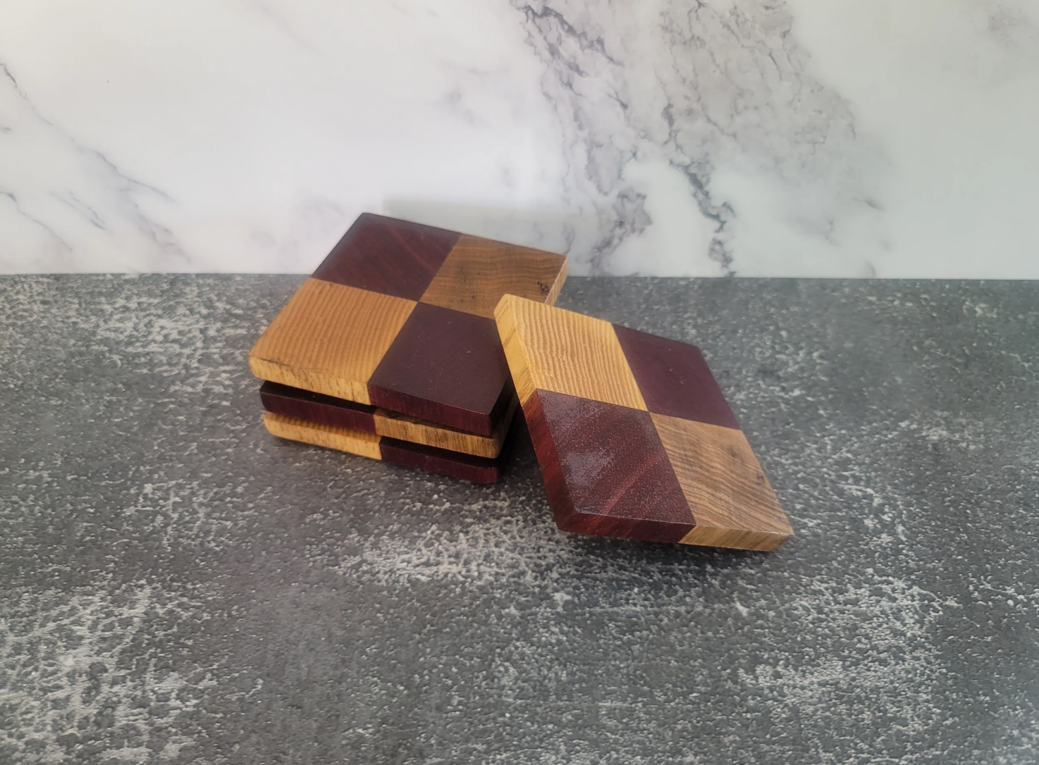 Stack of wooden checkerboard coasters with alternating light and dark squares on a dark speckled surface with a marble backsplash.