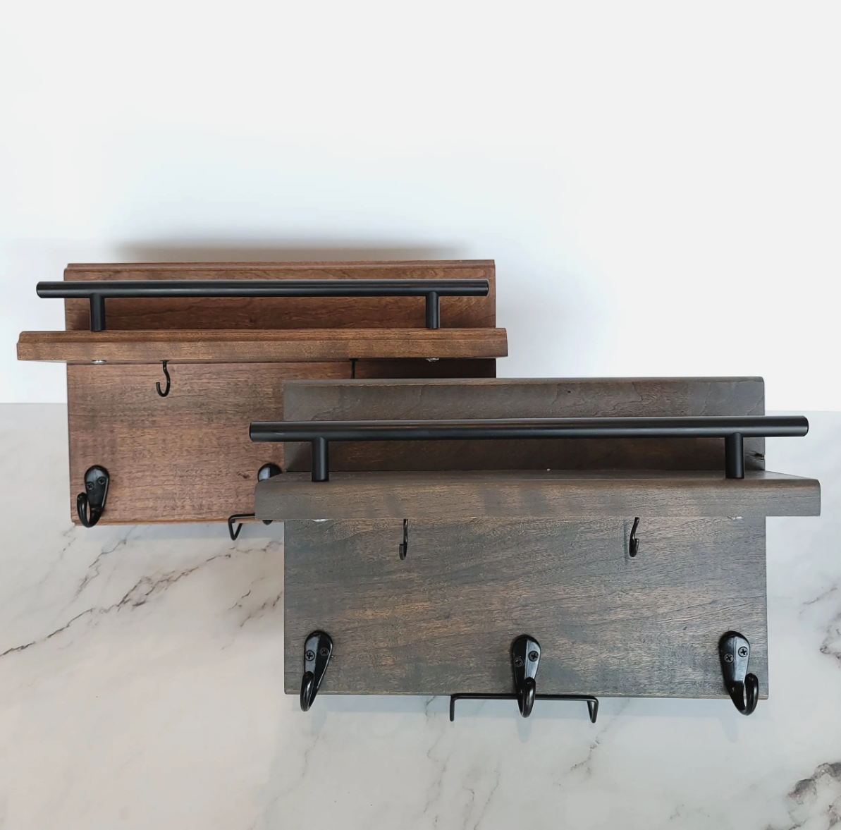 Two wooden wall-mounted coat racks with black metal hooks and handles, one in natural wood finish and the other in dark gray, on a white wall above a marble surface.