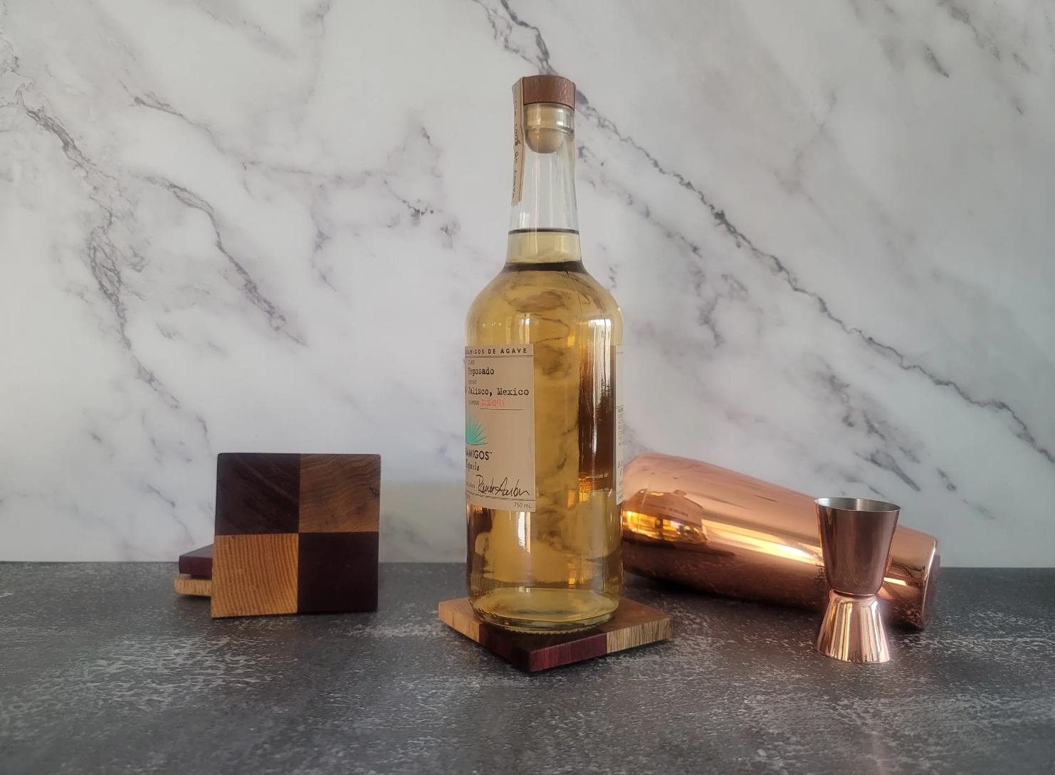 A bottle of agave spirit on a dark countertop with a marbled white background, accompanied by assorted barware and wooden blocks.