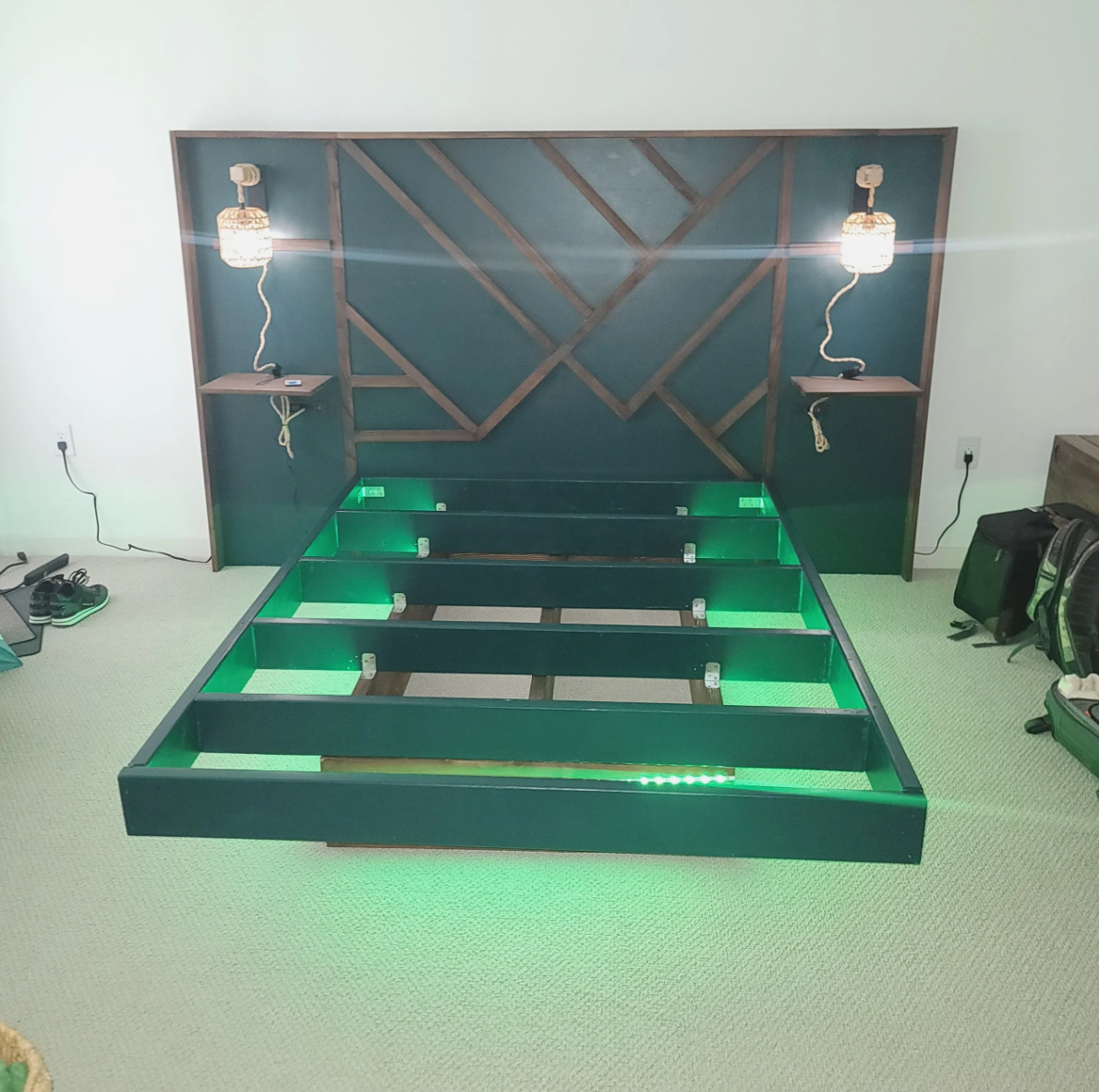 Empty green bed frame with LED lighting, headboard with geometric design, wall-mounted lamps on each side, and small shelves with a light switch, in a bedroom.