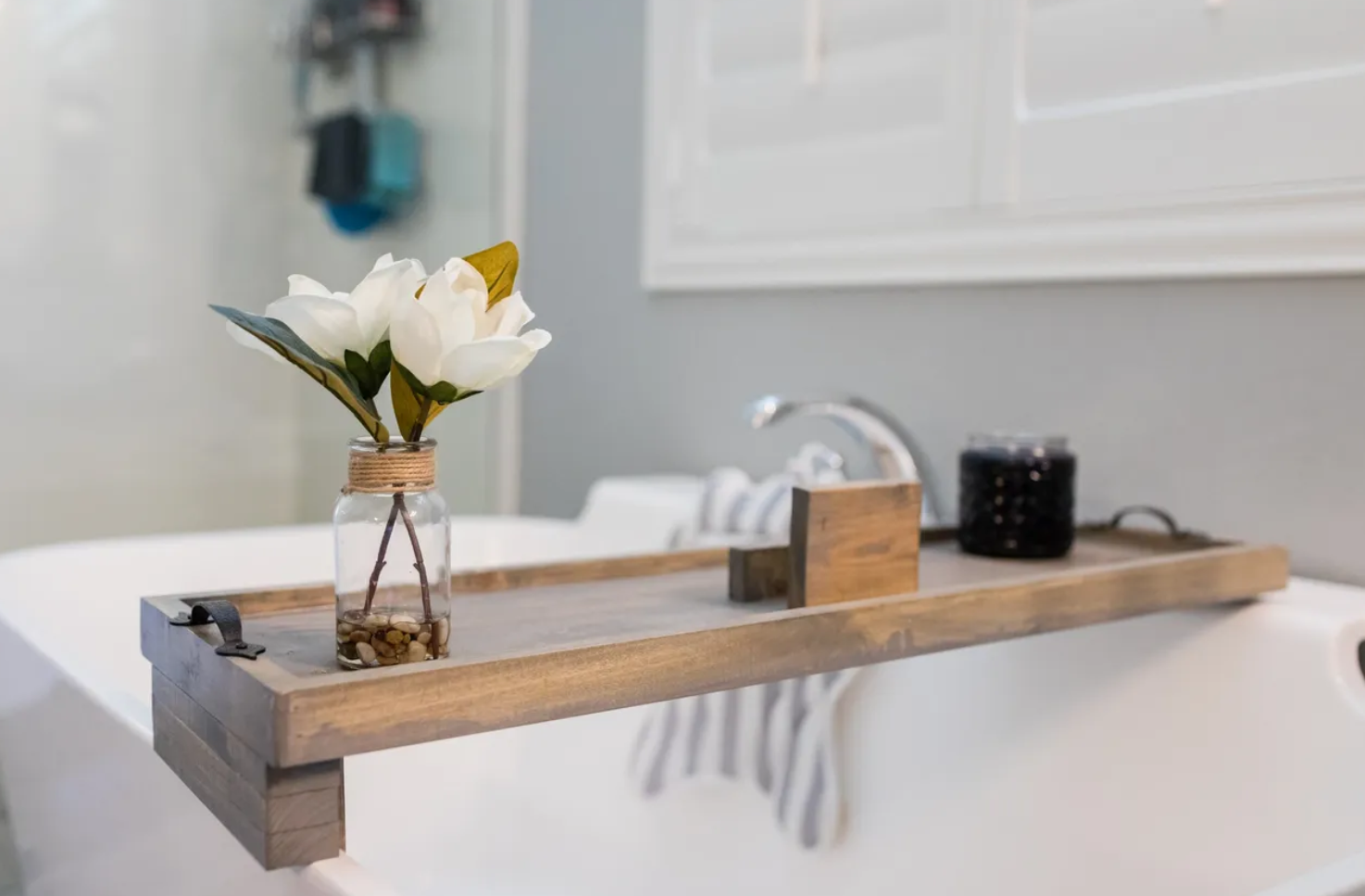 Decorative tray on a bathtub with a small glass jar containing white artificial flowers, a wooden soap holder, and a black container.