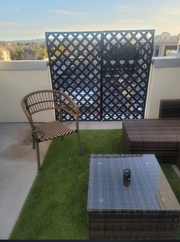An outdoor balcony with a wicker chair, a glass-topped wicker table with a small black object on it, a woven lounge chair, a decorative black lattice privacy screen, and a view of a cityscape and sky in the background.