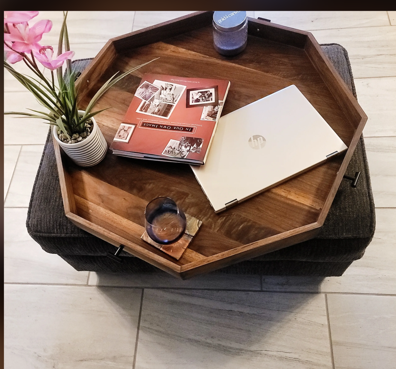 A wooden hexagonal tray on top of a dark upholstered ottoman containing a potted pink flower, a glass of water on a coaster, an open HP laptop, a brochure or magazine, and a bottle of blue-capped water.