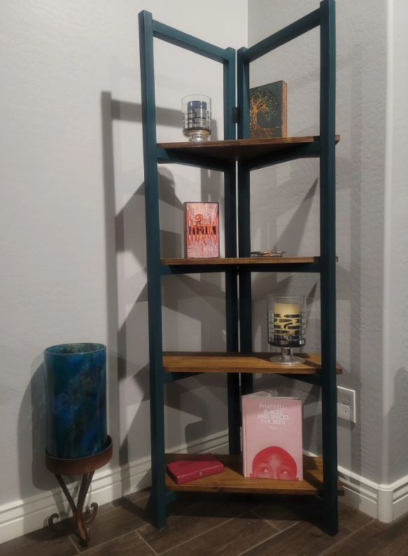 A tall wooden bookshelf with five shelves. The top shelf has a glass container and a framed picture of a tree. The second shelf has a red book and small objects. The middle shelf has a clear candle holder with a yellow candle and black-cutout letters