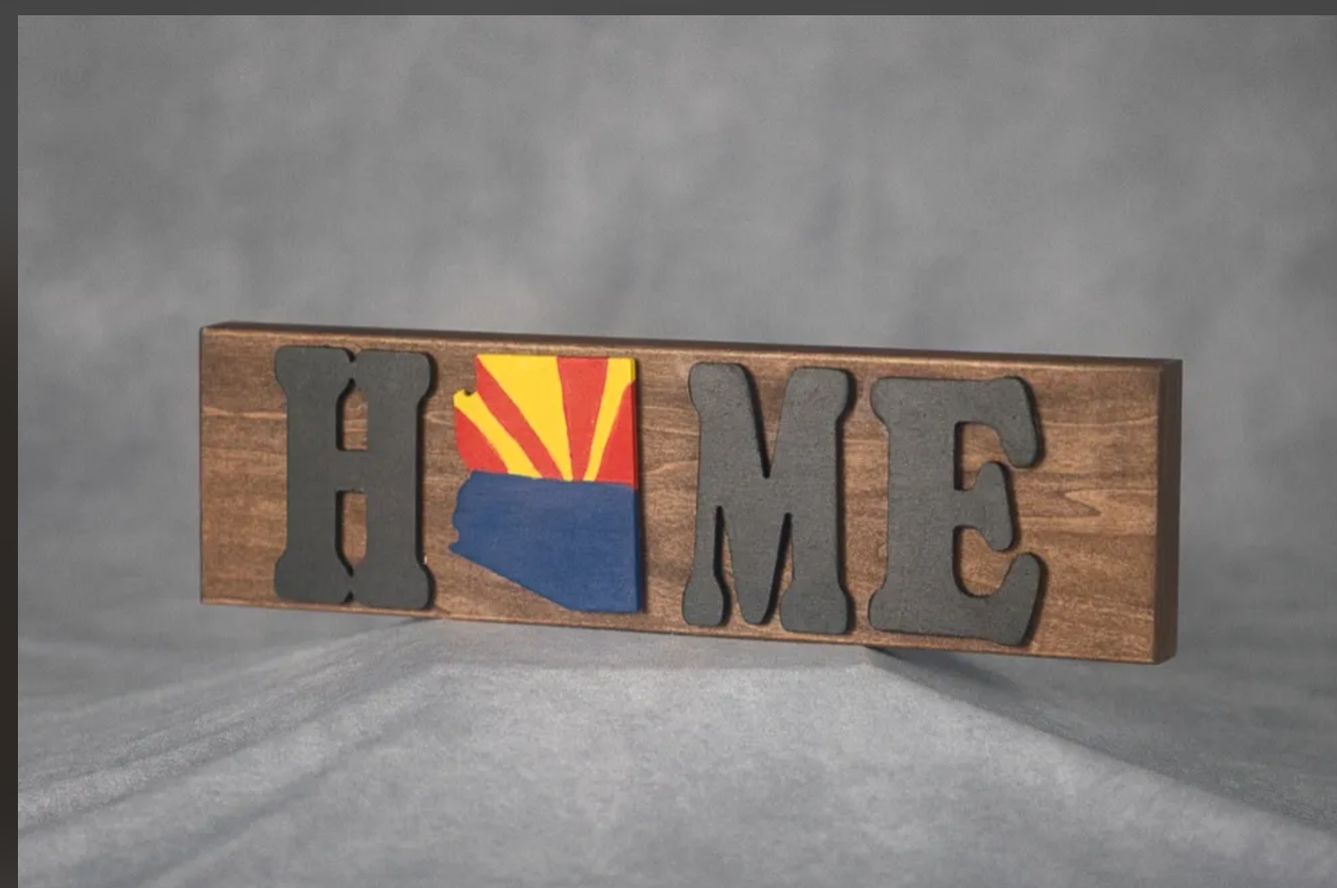 Wooden decorative sign spelling 'HOME' with the 'O' replaced by a painted Arizona state outline featuring a sunset and desert scene.