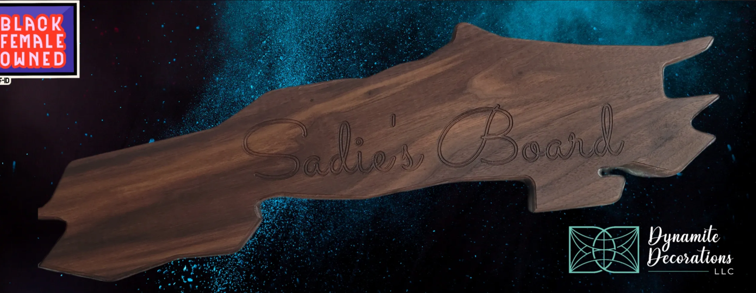 A wooden plaque shaped like the state of North Carolina with the inscription 'Sadie's Board' engraved on it. The background features a cosmic or galaxy-like pattern with a blue-speckled design. There is a logo in the bottom right corner that says 'Dy
