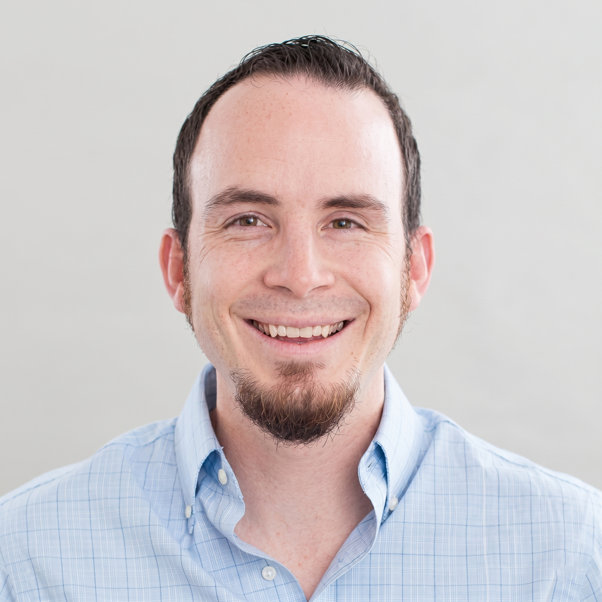 Close-up of a smiling man with short dark hair, light skin, and wearing a light blue checkered shirt.