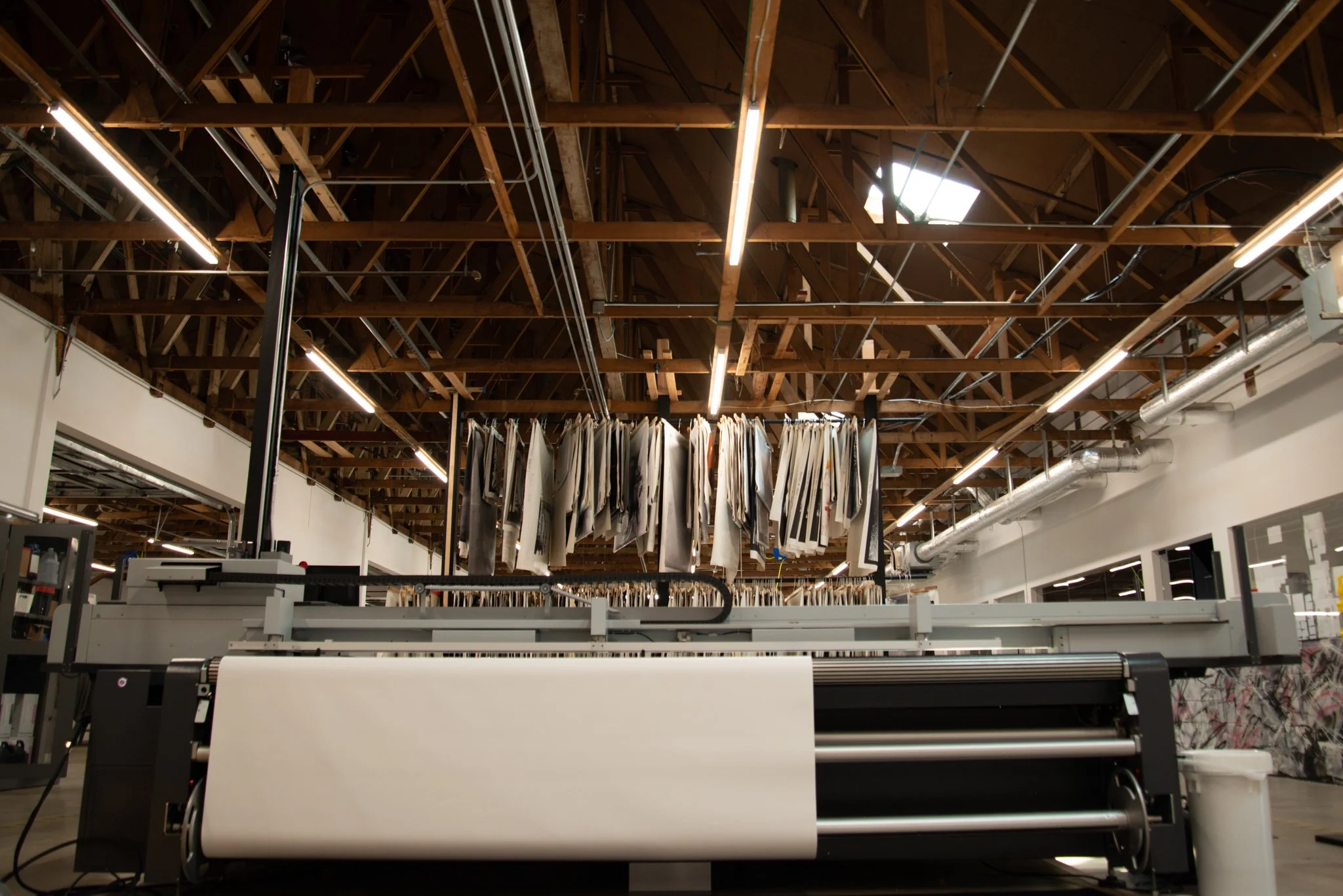 Interior of General Public's manufacturing facility showing a large printer with rolled canvases hanging from the ceiling, and bright lighting fixtures.