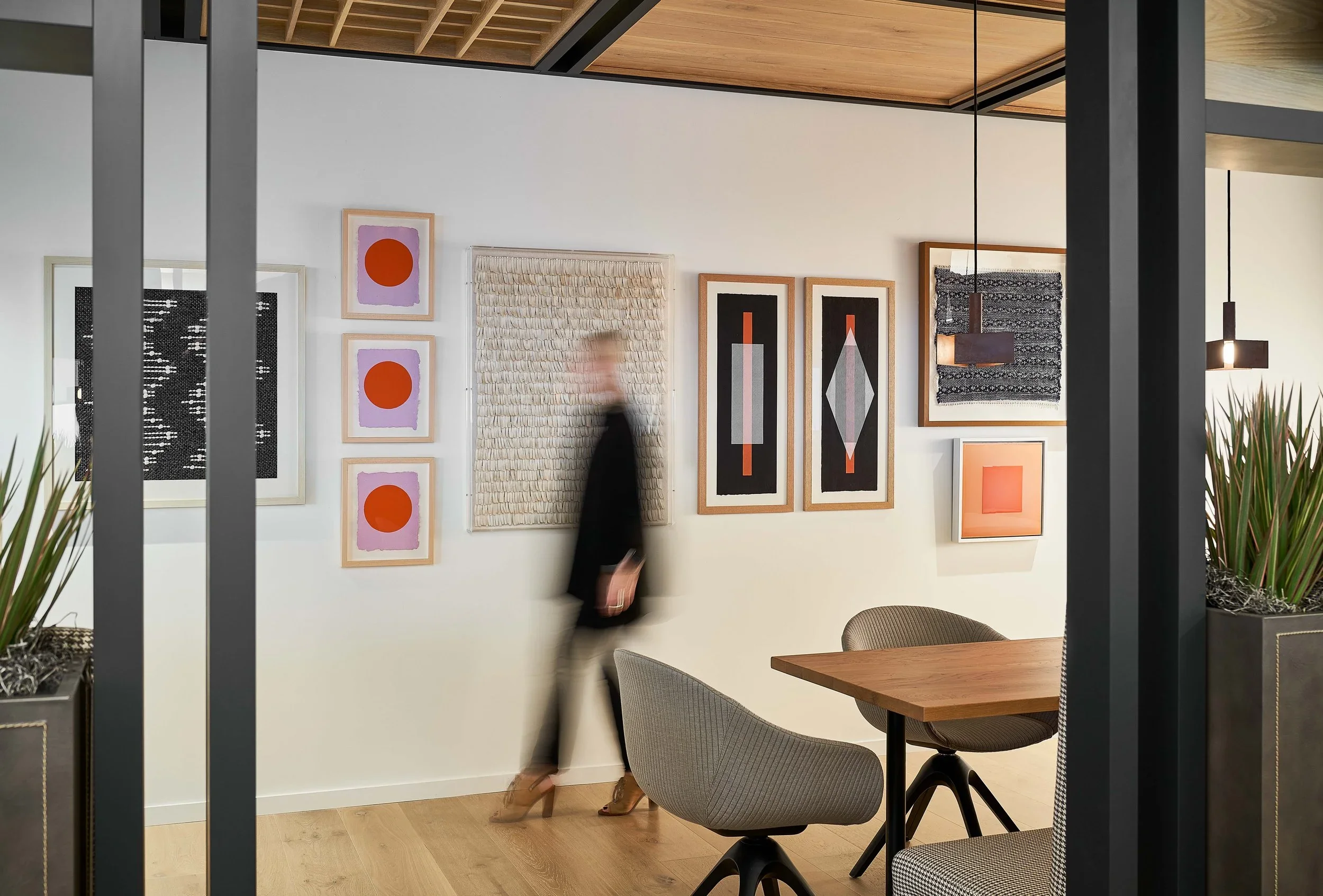 Interior of a commercial office with Simply Framed, General Public art replicas on a white wall, a blurred person walking, and seating area with gray chairs and wooden table.