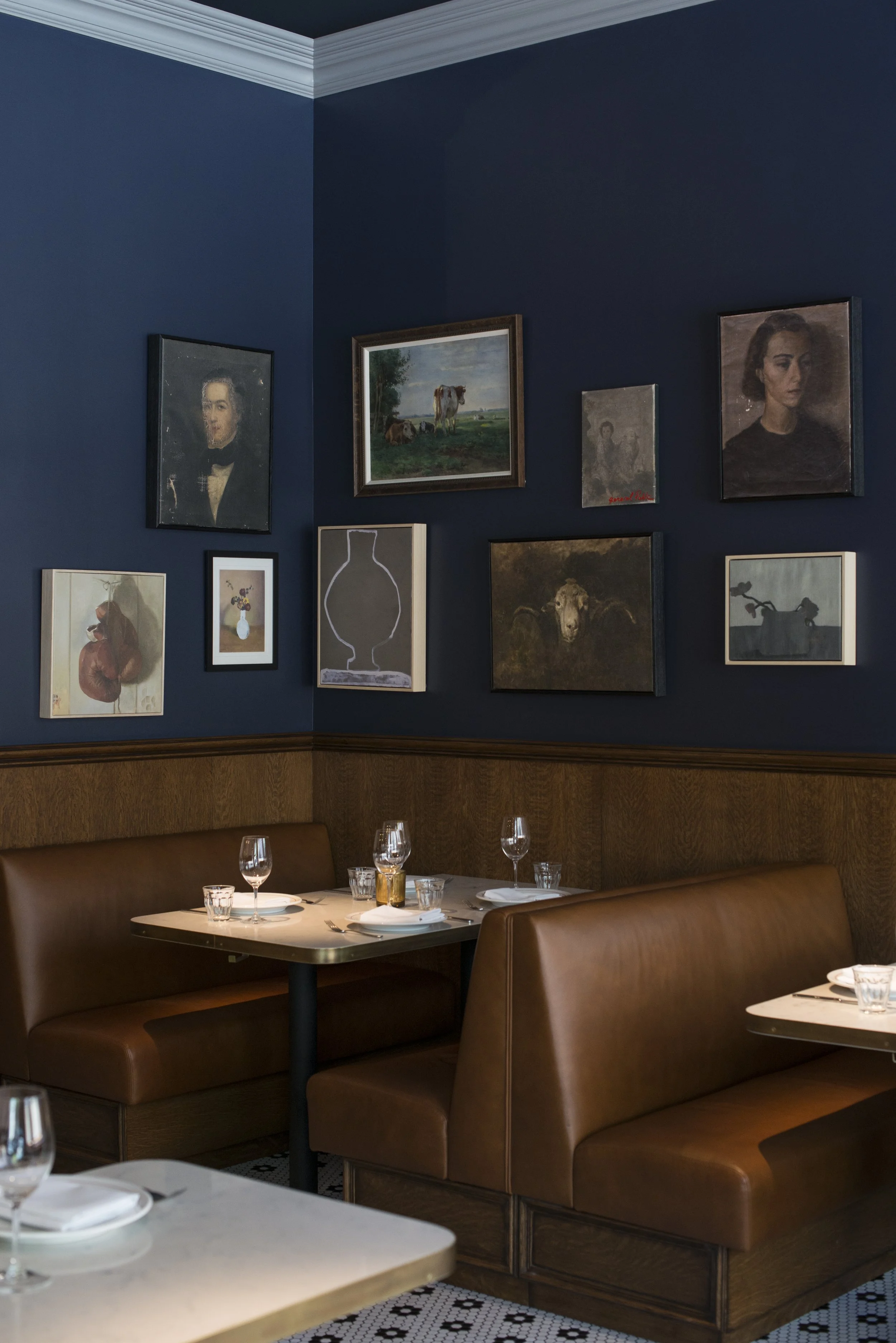 A corner of a restaurant dining area with tables set with glasses, napkins, and plates, brown leather booths, and a dark blue wall decorated with various framed paintings, including portraits and a landscape with cows.