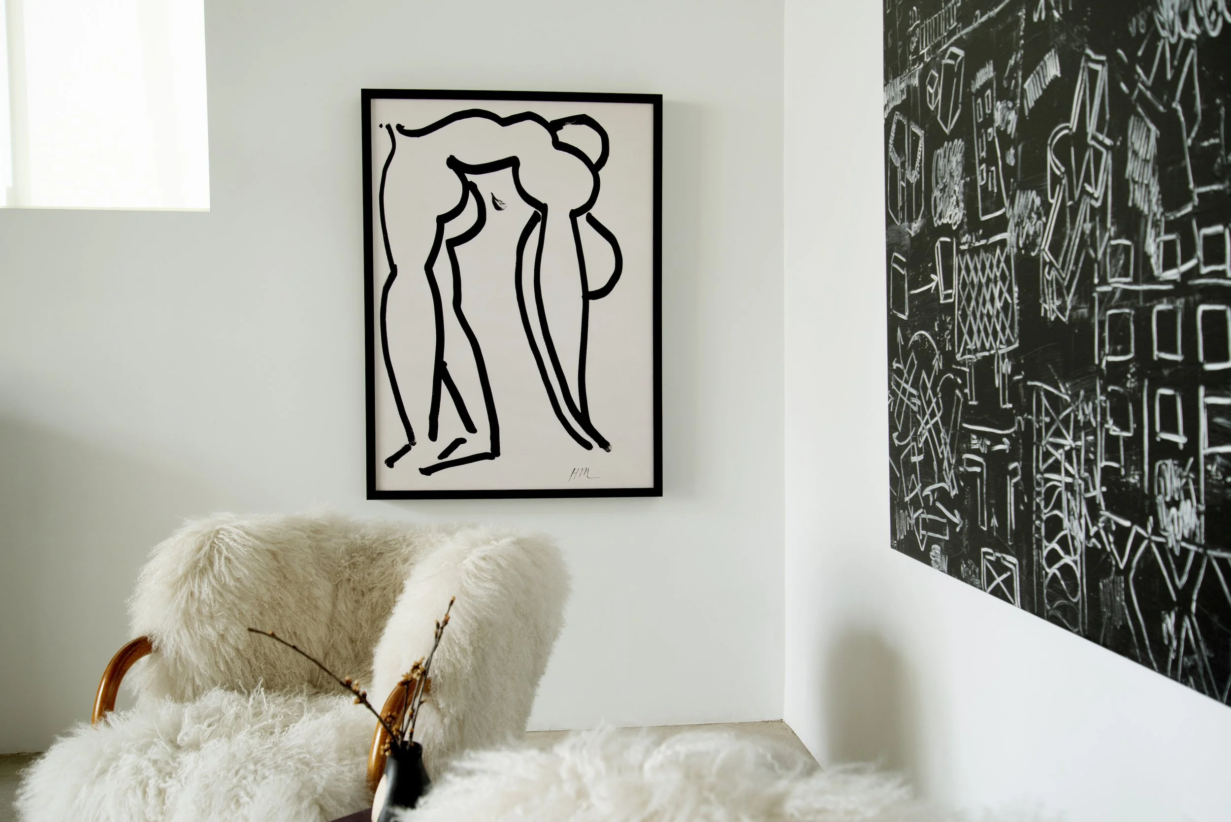 Interior view of a room with a white wall, a large framed black and white abstract line art, a black abstract artwork with white line drawings, a fluffy white sheepskin rug, and a chair with a sheepskin cover and wooden armrests.