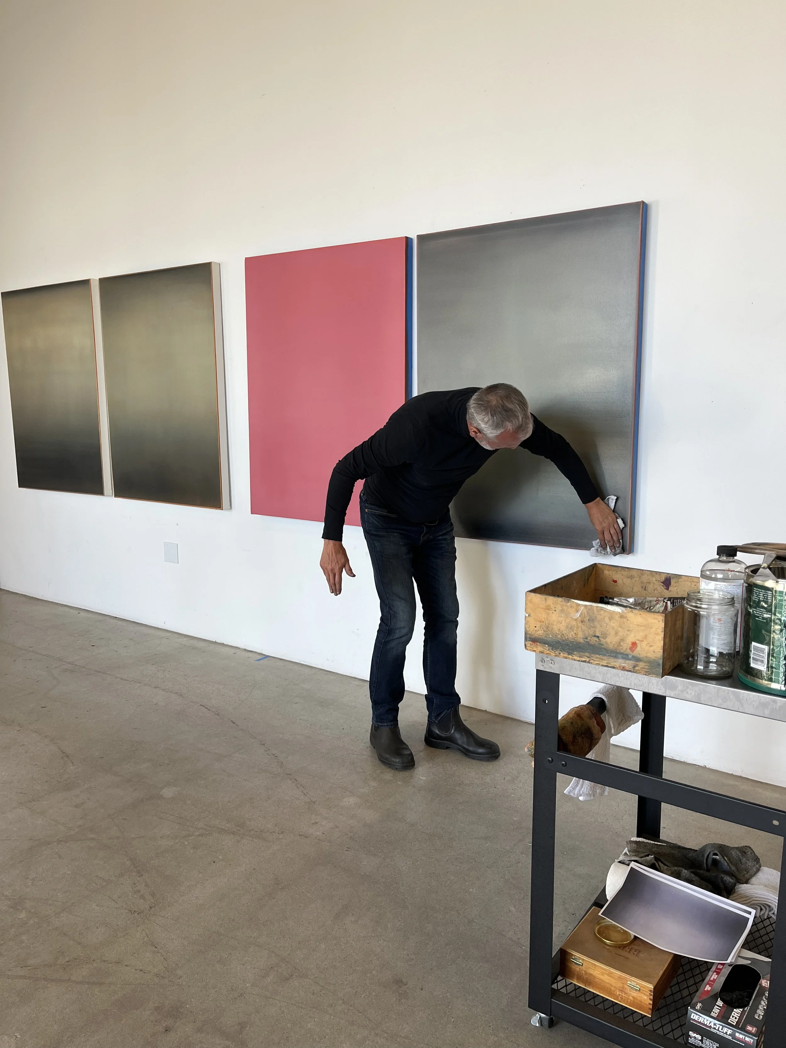 An artist painting a portrait that's hanging in a row on a wall in his gallery, in black shirt and jeans with art supplies on a table next to him. 