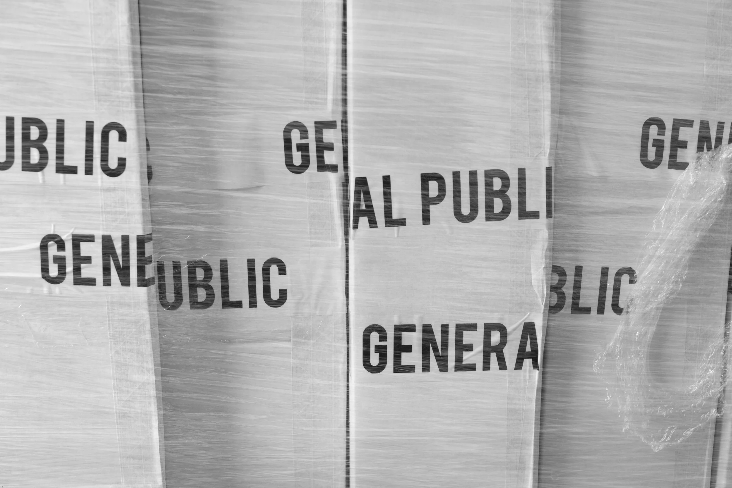 Black and white photo of multiple stacked cardboard boxes wrapped in plastic, with the words 'GENERAL' and 'PUBLIC' printed on them.