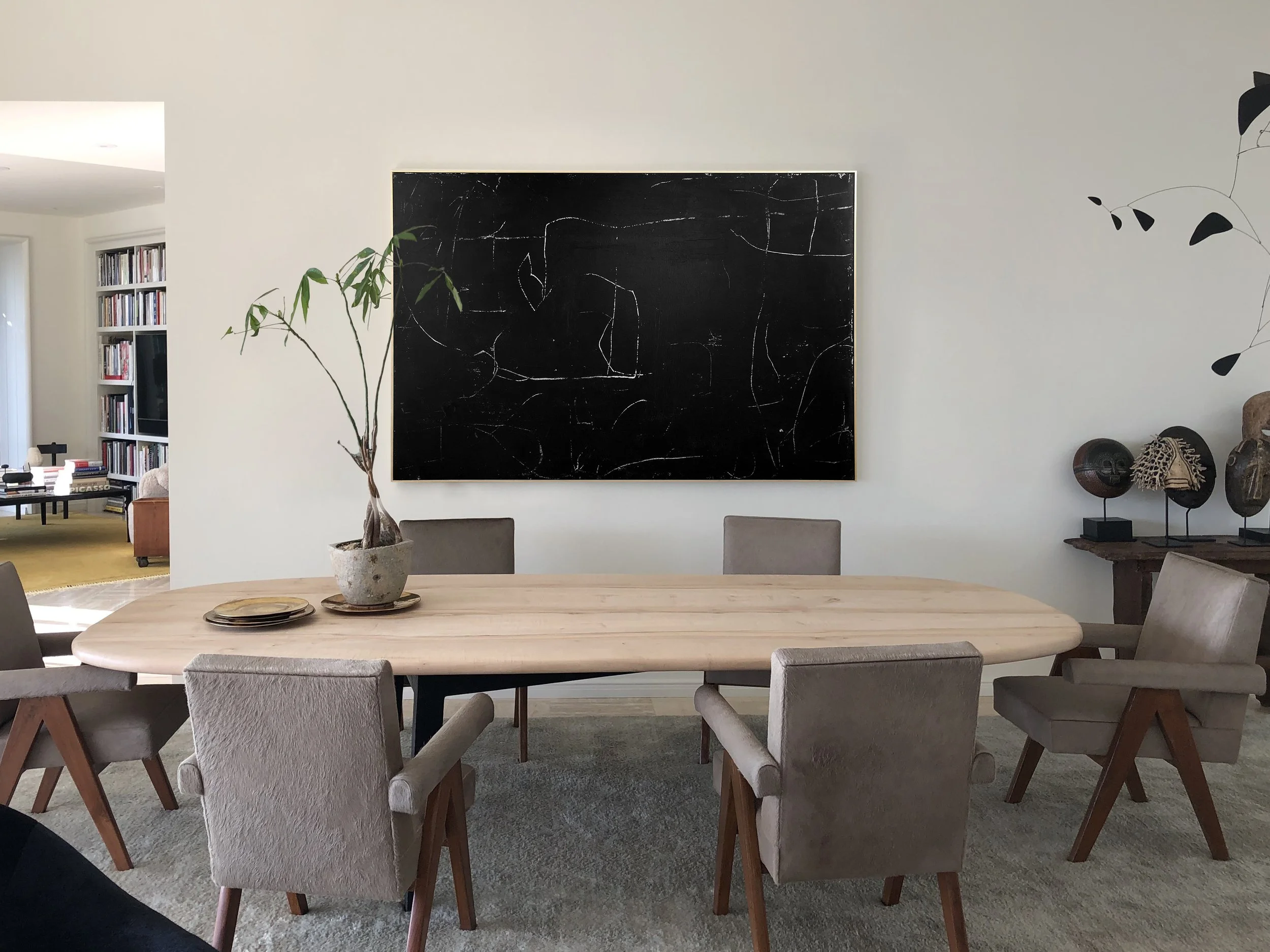 A room with a round wooden dining table surrounded by light gray upholstered chairs with wooden legs. A large black abstract painting with white lines hangs on an off-white wall. To the right, there are several African tribal masks on display. To the