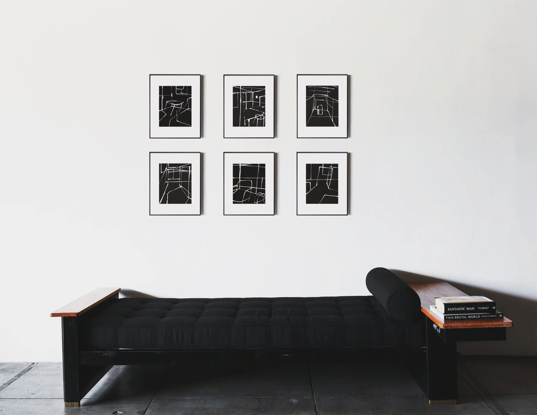 A black bench with a tufted seat against a white wall, with six, Simply Framed black and white line art drawing prints arranged in two rows hanging above it. To the right, a small wooden side table holds a stack of books.