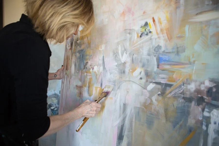 An artist with blond hair wearing a black shirt painting an abstract artwork on a large canvas with various colors including white, beige, pink, blue, and yellow.