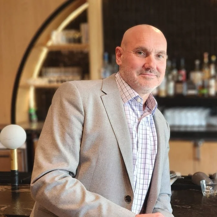 A middle-aged man with a bald head and gray beard in a bar or restaurant, wearing a light gray blazer and a checked shirt, standing near a bar counter with a background of liquor bottles.