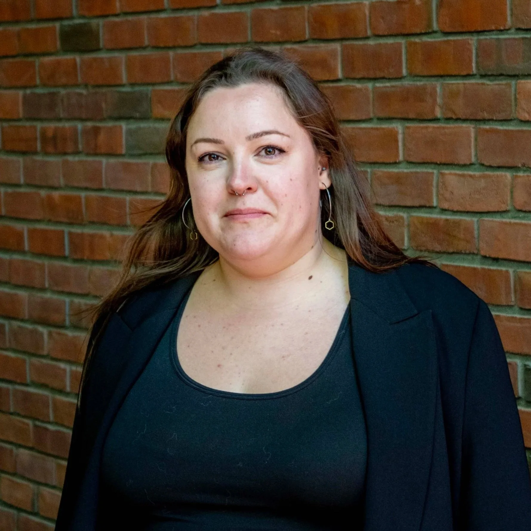 A woman with long brown hair, wearing a black top and earrings, standing against a brick wall.
