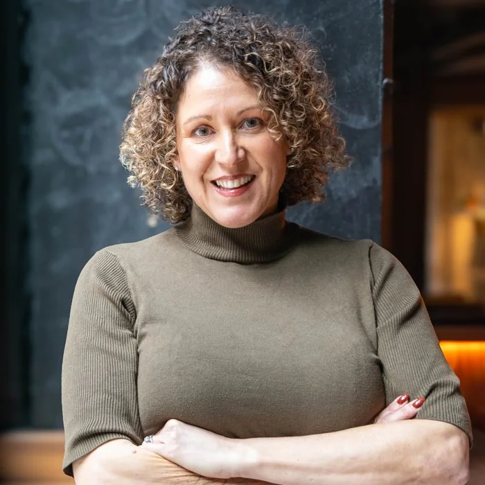 A woman with curly hair wearing a green turtleneck sweater, smiling with arms crossed, standing against a dark textured background.