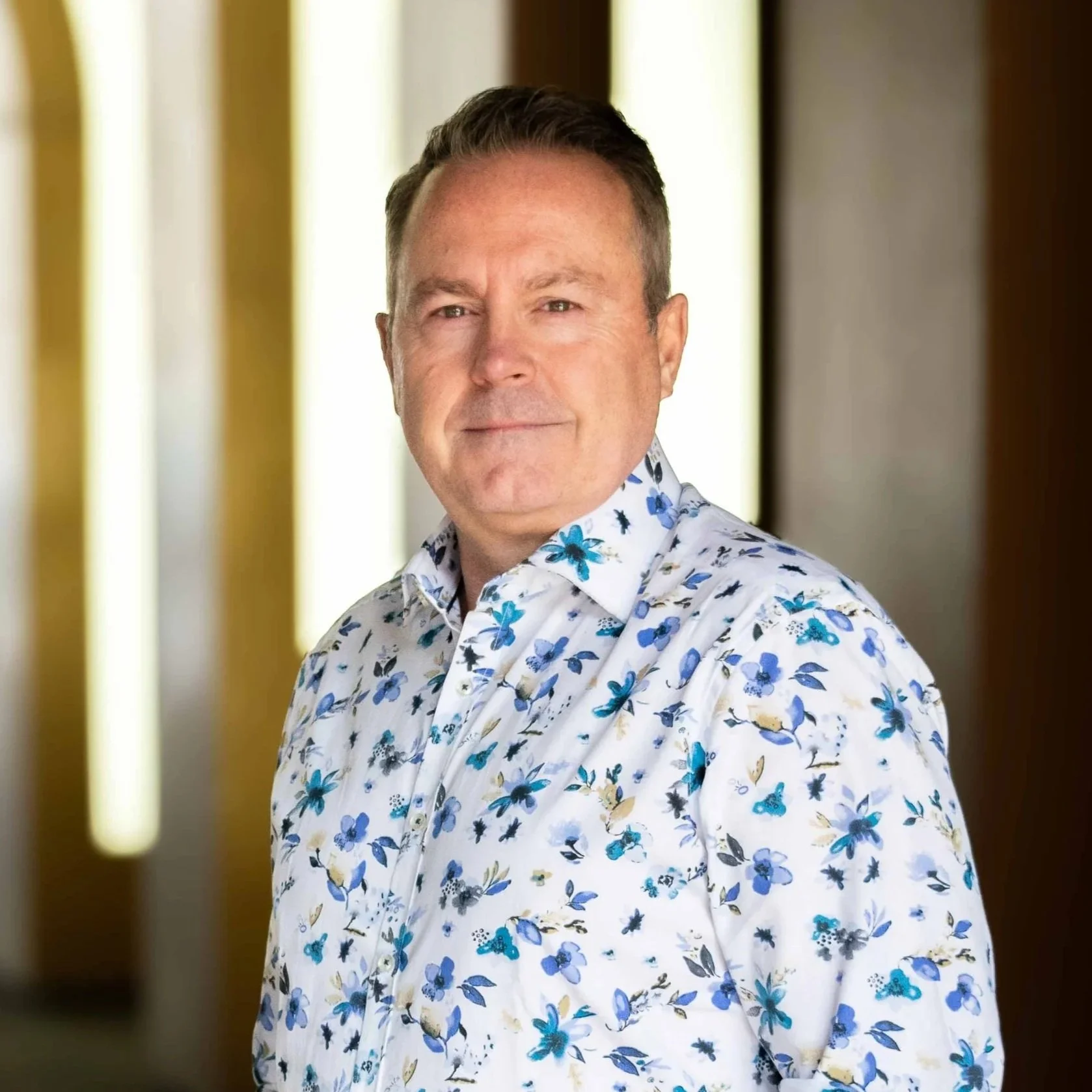 A middle-aged man with short brown hair and light skin, wearing a white shirt with a blue floral pattern, standing indoors with a blurred background including vertical light panels.
