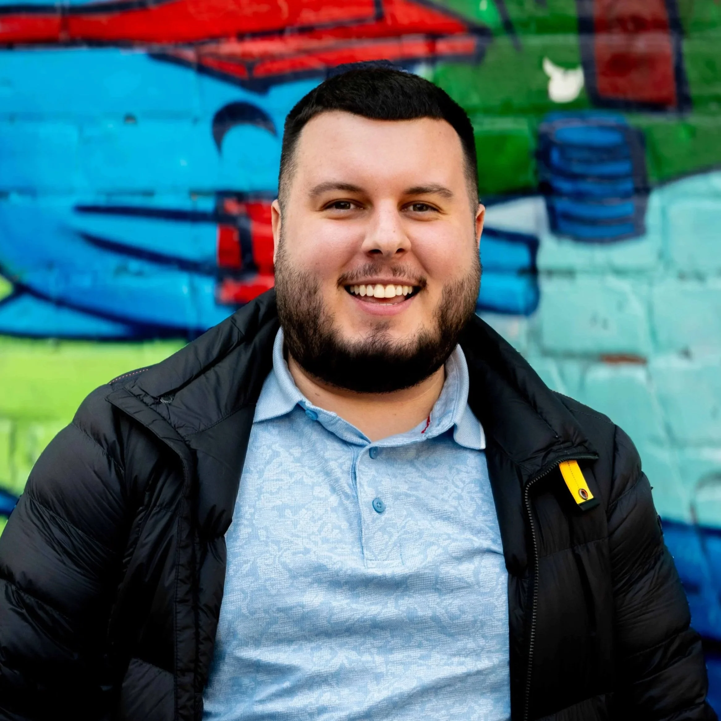 Smiling man with short dark hair and beard, wearing a dark jacket and light blue button-up shirt, standing in front of a colorful abstract mural.