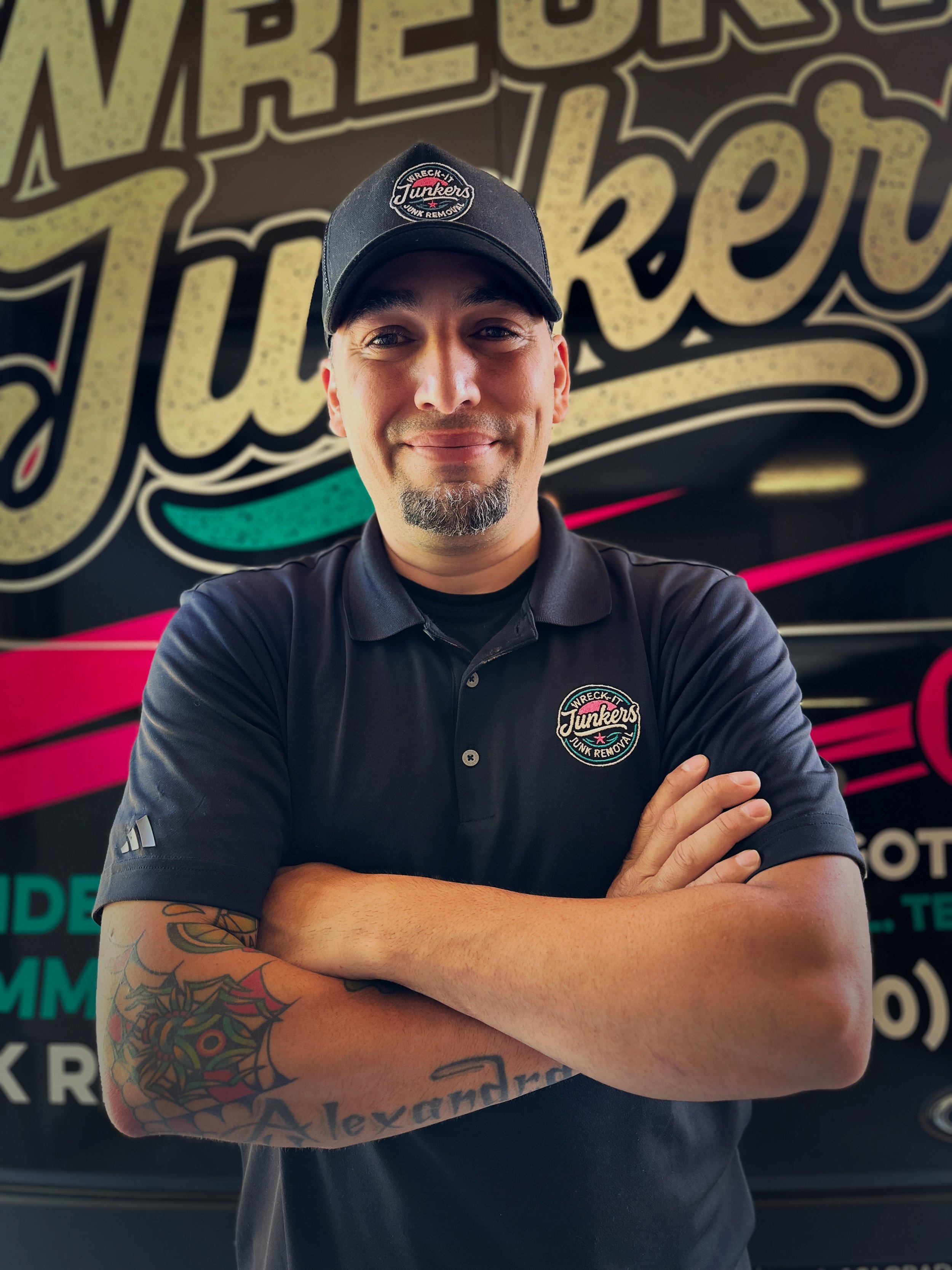 A smiling man wearing a black polo shirt and a black baseball cap with a logo, standing with arms crossed in front of a colorful background with large lettered text.