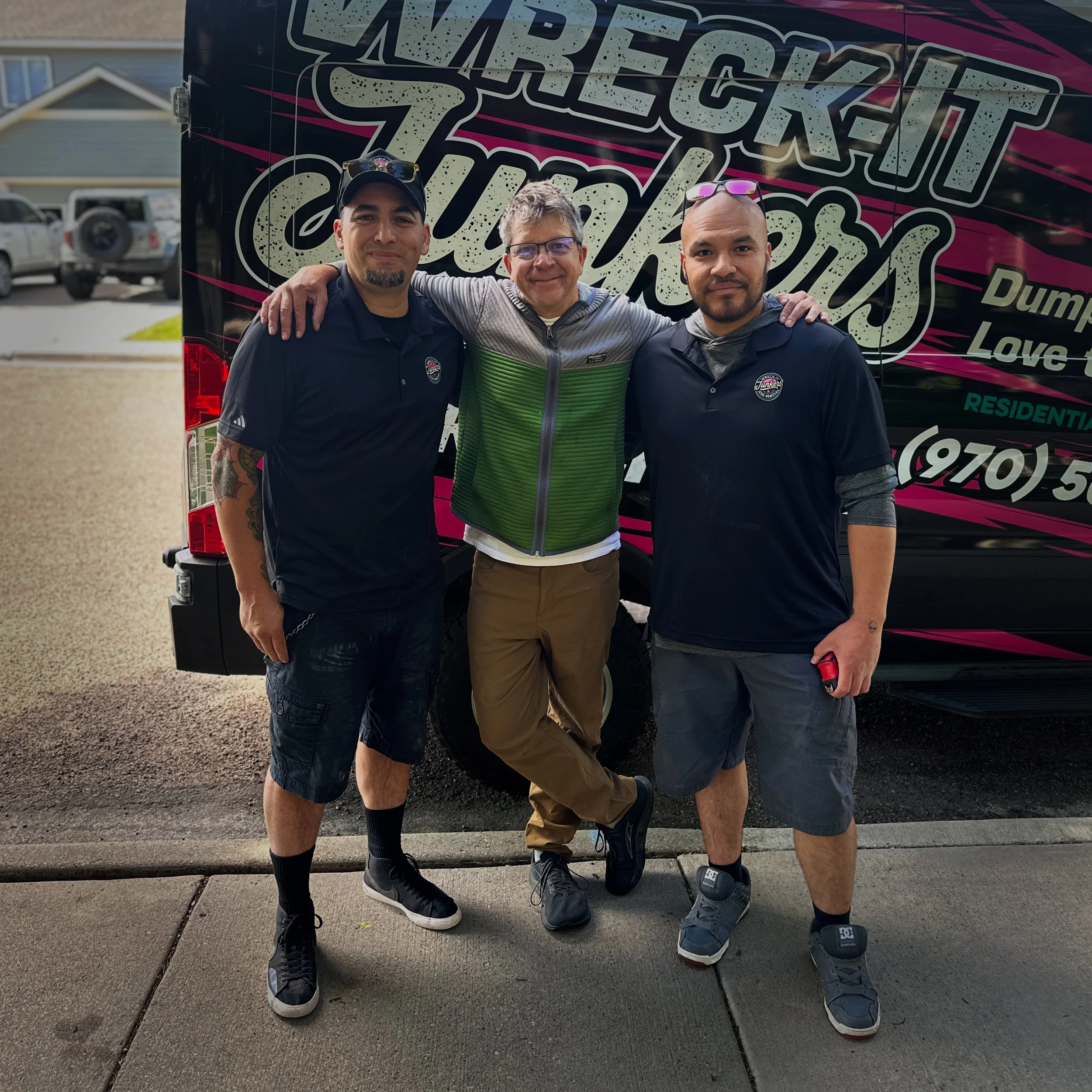 Three men standing close together outdoors in front of a large vehicle with bold graphic text, with houses and parked cars in the background. The men have their arms around each other, smiling at the camera.