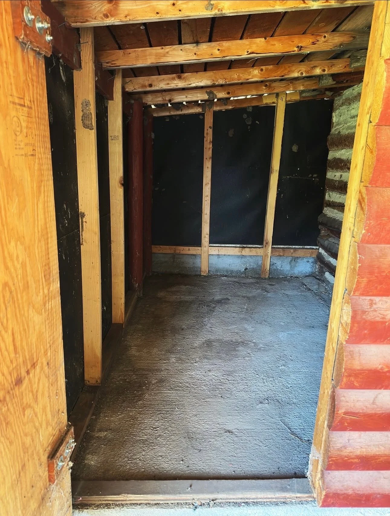 Interior view of a small unfinished room with exposed wooden beams and black sheathing on the walls, concrete floor, and partial brick wall on the right side.